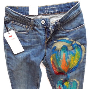 Peut inclure: Un jean bleu denim avec un motif floral abstrait coloré peint sur la jambe droite. Le jean a une coupe droite et une étiquette qui dit "BOLD CURVE 27 straight leg".