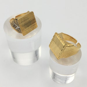 Könnte beinhalten: Zwei goldene Ringe mit quadratischen Oberseiten. Die Ringe werden auf klaren Acrylständern präsentiert.