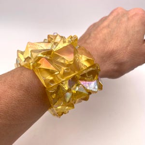 Peut inclure: Un bracelet en cristal jaune iridescent composé de verre facetté. Le bracelet est composé de nombreuses petites pièces de verre disposées en grappe. Le bracelet est porté au poignet.