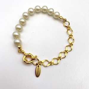 Peut inclure: Un bracelet en chaîne dorée avec une rangée de perles blanches. Le bracelet a un fermoir avec le mot "KIKINASI" dessus.