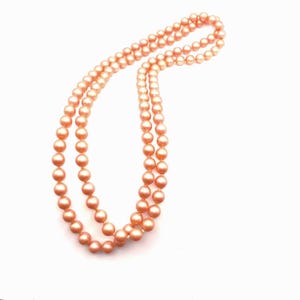 May include: A double strand necklace of peach-coloured, round beads. The necklace is set against a white backdrop, with the strands elegantly draped. The beads appear uniform in size and have a subtle sheen, suggesting a classic jewellery design.