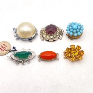 Peut inclure: Un ensemble de six fermoirs à bijoux décoratifs présentant divers designs : une grosse perle artificielle crème, une pierre rouge ronde avec des cristaux clairs, un groupe de petites perles bleues, une pierre marquise verte, un cabochon orange ovale et un cristal floral jaune.