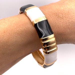 Peut inclure: Un bracelet jonc doré avec des sections alternées noires, blanches et dorées. Le bracelet a une finition brillante et est porté au poignet. Le design présente un motif géométrique audacieux.