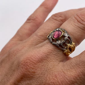 Può includere: Un anello con una pietra cabochon ovale rosa scuro al centro, incastonata in una montatura dorata. La fascia è realizzata in metallo color argento scuro ossidato con un motivo a filigrana traforato, arricchito da piccole pietre chiare.