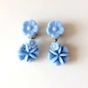 Peut inclure: Une paire de boucles d'oreilles en forme de fleurs en verre bleu clair. Les boucles d'oreilles sont composées de plusieurs petites fleurs regroupées. La fleur du haut est plus grande et a un seul centre.