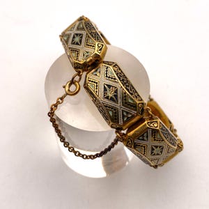 Peut inclure: Un bracelet doré avec des maillons géométriques et facettés. Chaque maillon présente des détails complexes en émail noir, argent et sarcelle. Une petite chaîne sécurise le bracelet. Le bracelet est présenté sur une surface claire et arrondie.