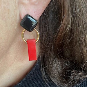 Peut inclure: Boucle d'oreille unique avec un carré noir, un anneau doré et un pendentif rectangulaire rouge. La boucle d'oreille est portée sur une oreille, avec les cheveux et l'épaule de la personne visibles en arrière-plan.