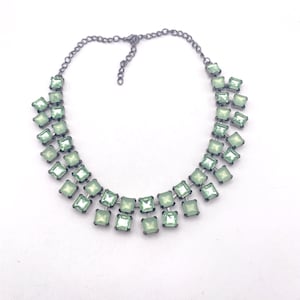 Può includere: Una collana statement con due file di pietre quadrate sfaccettate color verde menta chiaro, incastonate in montature di metallo grigio scuro. La collana è collegata da una catena in metallo scuro con chiusura a moschettone e catenella.