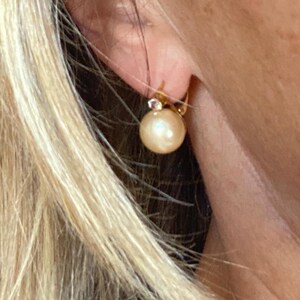 Peut inclure: Gros plan d'une boucle d'oreille dorée avec une grosse perle ronde de couleur crème. La boucle d'oreille a un petit accent de pierre précieuse transparente au-dessus de la perle. La boucle d'oreille est attachée à un crochet doré.