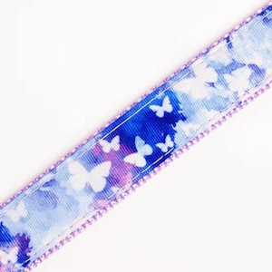 May include: A close-up of a patterned ribbon featuring a butterfly design. The ribbon has a blue and purple watercolor background with white butterfly silhouettes. The edges are trimmed with a lavender stitching.