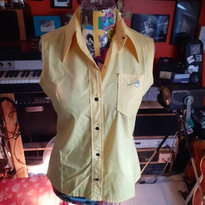 May include: A sleeveless yellow button-down shirt with a blue embroidered logo on the left chest pocket.