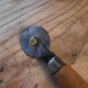 May include: A vintage rotary tool with a circular blade and a wooden handle. The blade is made of metal and has a brass centre. The handle is made of wood and has a metal cap.
