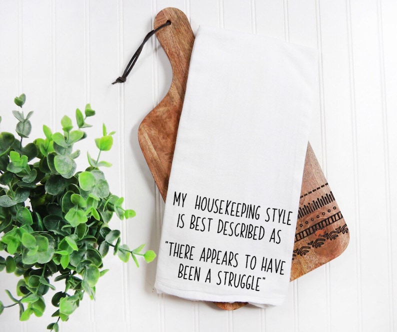 May include: White kitchen towel with black text that reads "My housekeeping style is best described as "There appears to have been a struggle."
