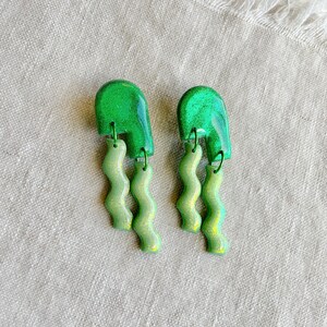May include: A pair of green and gold glitter earrings with a wavy design. The earrings are made of polymer clay and have a unique, modern look.