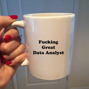 Op de afbeelding: Witte keramische mok met de tekst "Fucking Great Data Analyst" in het zwart. De mok wordt vastgehouden door een hand met rode nagellak.