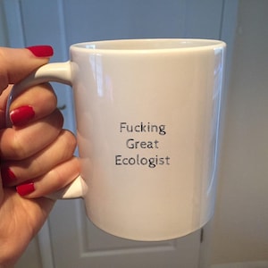 Può includere: Tazza in ceramica bianca con manico curvo. La tazza presenta la scritta "Fucking Great Ecologist" in nero, centrata sul davanti. La tazza è tenuta da una mano con lo smalto rosso.