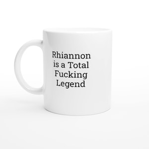 Puede incluir: Taza de cerámica blanca con asa curva. La taza presenta el texto "Rhiannon is a Total Fucking Legend" en negro, centrado en la parte delantera. La taza está sobre un fondo blanco.