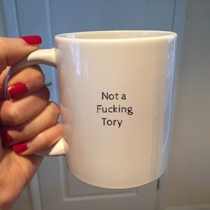 May include: White ceramic mug with the text "Not a Fucking Tory" in black. The mug has a white handle. The mug is held by a hand with red nail polish.