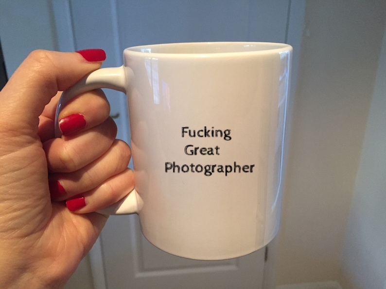 The hands of a person holding a white 11oz ceramic mug with the words Fucking Great Photographer printed on it. The text on the mug is grey and grainy.