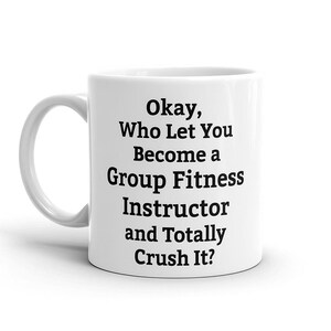 May include: White ceramic mug with a curved handle. The mug features black text that reads: "Okay, Who Let You Become a Group Fitness Instructor and Totally Crush It?" A humorous gift for fitness instructors.