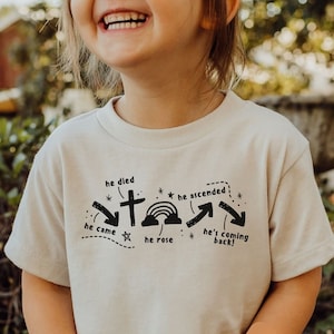May include: A light beige toddler t-shirt with a black graphic design featuring a cross, stars, a rainbow, and arrows. The text on the shirt reads "he died", "he came", "he rose", "he ascended", and "he's coming back!"