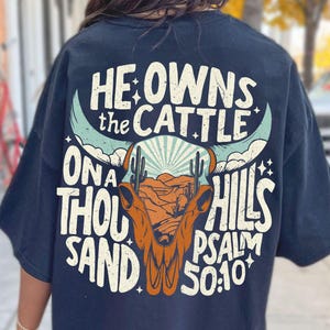 May include: Navy blue t-shirt with a graphic design on the back. The design features a bull skull, cacti, and the text "HE OWNS THE CATTLE ON A THOUSAND HILLS PSALM 50:10". The shirt is a casual wear item.