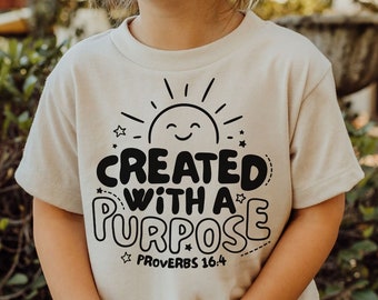 May include: A light beige t-shirt with a black graphic that says "Created with a Purpose" and "Proverbs 16:4". The graphic also includes a sun and stars.