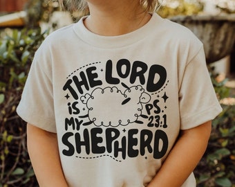 May include: A child wearing a beige t-shirt with a black graphic that says "The Lord is my Shepherd" with a sheep illustration and the text "Ps. 23:1".