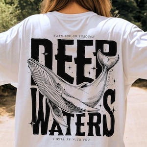 Pode incluir: Camiseta branca com um gráfico preto de uma baleia e as palavras "DEEP WATERS" e "I WILL BE WITH YOU". A camiseta também tem o texto "WHEN YOU GO THROUGH" e "ISAIAH 43:2".