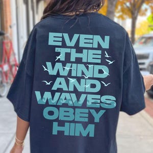 May include: A navy blue oversized t-shirt with the text "EVEN THE WIND AND WAVES OBEY HIM" in teal. The text is arranged in a stacked format with stylized wave and bird graphics. The shirt is worn outdoors.