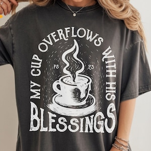 May include: Dark gray t-shirt with a white graphic. The graphic features a coffee cup with steam and the text "MY CUP OVERFLOWS WITH HIS BLESSINGS" in a circular design. The shirt has short sleeves and a relaxed fit.