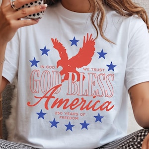 May include: A white t-shirt featuring a red eagle silhouette surrounded by a circle of blue stars. The text reads: IN GOD WE TRUST, GOD BLESS America, 250 YEARS OF FREEDOM. The design uses red and blue ink on a plain white short-sleeved cotton shirt.