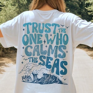 以下が含まれることがあります： 背中に青とターコイズ色のグラフィックが入った白いTシャツ。「TRUST THE ONE WHO CALMS THE SEAS」の文字と波のイラスト、聖書のマルコ4:35-41の聖句が描かれています。