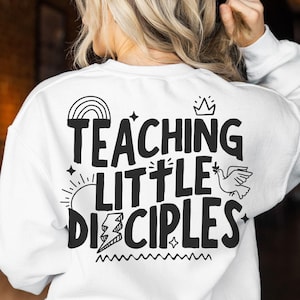 Könnte beinhalten: Weißer Pullover mit schwarzem Text, der "Teaching Little Disciples" lautet, mit einem Regenbogen, einer Krone, einer Taube und einer Sonne.