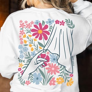 Puede incluir: Sudadera blanca con un diseño floral en la espalda. El diseño presenta una mano que atraviesa una cortina de tela para tocar una flor rosa, rodeada de flores y vegetación coloridas. El texto en el lado derecho dice "MATT 6:28-32".
