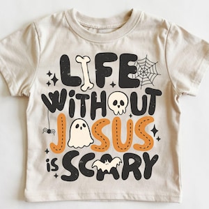 May include: A beige short-sleeve t-shirt featuring black and orange graphic text that reads "LIFE WITHOUT JESUS IS SCARY." The design includes illustrations of a bone, a spider web, a skull, a ghost, a small spider, and a bat.