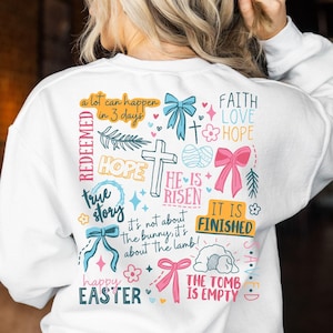May include: White sweatshirt with a colourful Easter design featuring the words "a lot can happen in 3 days", "REDEEMED", "HOPE", "true story", "HE IS RISEN", "IT IS FINISHED", "FAITH LOVE HOPE", "happy EASTER", and "THE TOMB IS EMPTY".