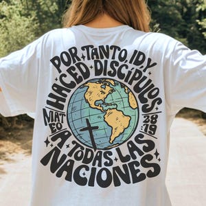 Könnte beinhalten: Weißes T-Shirt mit einem Aufdruck auf der Rückseite, der einen Globus mit gelben Kontinenten und einem Kreuz zeigt. Der Text lautet "POR TANTO, ID Y, HACED DISCIPULOS, MAT 28:19, A TODAS LAS NACIONES" in Schwarz und Blau.