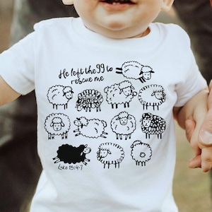 Op de afbeelding: Wit T-shirt met korte mouwen met een zwarte schapenillustratie en de tekst "He left the 99 to rescue me" en "Luke 15:4-7". Het T-shirt wordt door een volwassene vastgehouden.