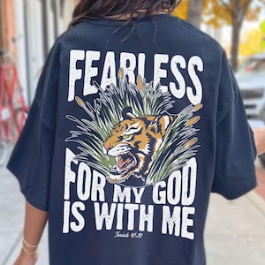 Może przedstawiać: Czarny, obszerny t-shirt z grafiką tygrysa i białym napisem "FEARLESS FOR MY GOD IS WITH ME". Tygrys jest pomarańczowo-czarny, na tle zielonych trzcin. Poniżej znajduje się tekst "Izajasza 41:10".