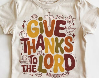 May include: A cream-colored short-sleeve t-shirt featuring a graphic print with the text "GIVE THANKS TO THE LORD" and "PS 107:1-3". The design includes autumn-themed illustrations like a pumpkin, a pie, leaves, a cross, and a dove.