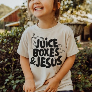 May include: A child wearing a light beige t-shirt with a black graphic that says "Juice Boxes & Jesus". The graphic includes a cartoon juice box with an apple inside.