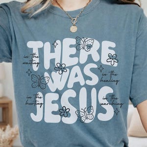 May include: A dusty blue t-shirt with the words "THERE WAS JESUS" in large white letters. The text is surrounded by small butterfly and flower illustrations. Additional text reads "in the waiting, in the healing, in the hurting, in the searching."