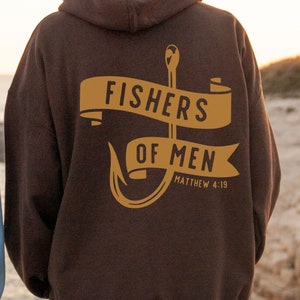 Puede incluir: Sudadera con capucha marrón con un gráfico dorado de un anzuelo de pesca y una pancarta que dice "Fishers of Men" con la referencia bíblica "Mateo 4:19".