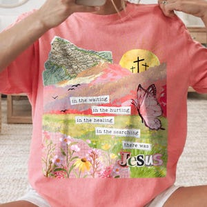 May include: A coral-colored t-shirt featuring a collage design with a butterfly, a yellow sun, and text that reads "in the waiting, in the hurting, in the healing, in the searching, there was Jesus."