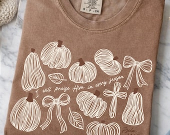May include: A brown short-sleeve t-shirt featuring a white line-art graphic of various pumpkins, two bows, a cross, and two leaves. The text reads, I will praise Him in every season, and Psalm 146:2. The shirt has a crew neckline and a visible brand label.