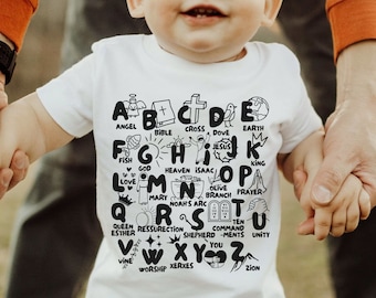 Puede incluir: Una camiseta blanca con un diseño de alfabeto negro. Cada letra está emparejada con una palabra, como "A" para "Ángel" y "B" para "Biblia".
