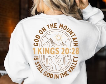 Könnte beinhalten: Weißer Pullover mit einem braunen Grafikdesign, auf dem "God on the Mountain 1 Kings 20:28 is still God in the Valley" steht.