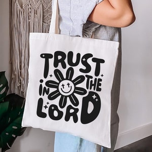 May include: A white canvas tote bag with the words "TRUST IN THE LORD" in large, black, retro-style lettering. The word "IN" is centered within a smiling flower design. The bag is held by a person.