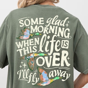 以下が含まれることがあります： オリーブグリーンのTシャツ。背中には3羽の鴨が飛ぶグラフィックと「SOME glad MORNING WHEN THIS life IS OVER I'll fly away」という文字がプリントされています。デザインには、かすれたオフホワイトのフォントと、カラフルな鴨のイラストが使用されています。